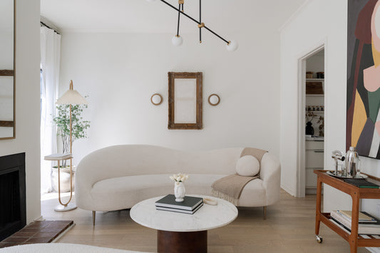 A minimalist living room with a curved cream sofa, a marble coffee table, and an abstract painting. The space is accented by a sculptural light fixture, natural greenery, and soft textures, creating a serene, modern aesthetic.