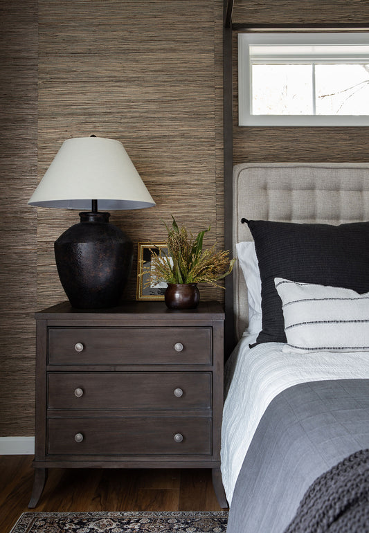 A cozy bedroom scene featuring a dark wood nightstand adorned with a rustic lamp, decorative vase, and framed artwork. The neutral tones, textured wallpaper, and soft bedding create a warm, inviting space, embodying timeless interior design elegance.