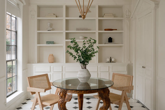 A sophisticated dining area featuring a glass table with carved wood legs, woven chairs, and minimalist decor. Elegant built-in shelves and natural light complement the greenery centerpiece, enhancing modern home decor.